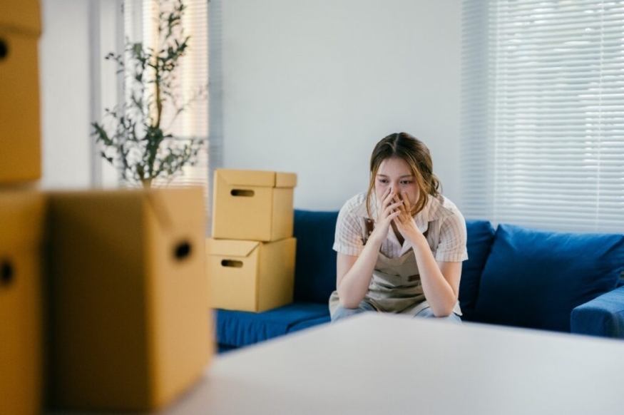A young woman sitting on a couch looks stressed while surrounded by moving boxes in a living room.