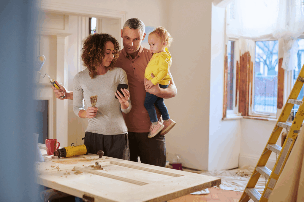 A couple with their young child standing in a room under renovation, looking at a smartphone together. The woman holds a paint scraper, while the man holds the child in one arm. A worktable with tools, a ladder, and partially stripped window frames are visible in the background.