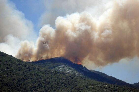 Large wildfire producing heavy smoke over a mountain landscape, with an aircraft assisting in fire monitoring or suppression.