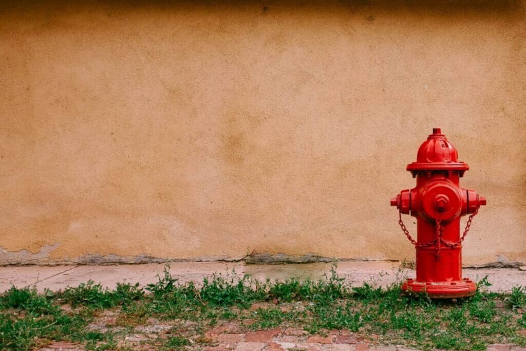 Red fire hydrant positioned beside a beige exterior wall with grass and pavement in the foreground.
