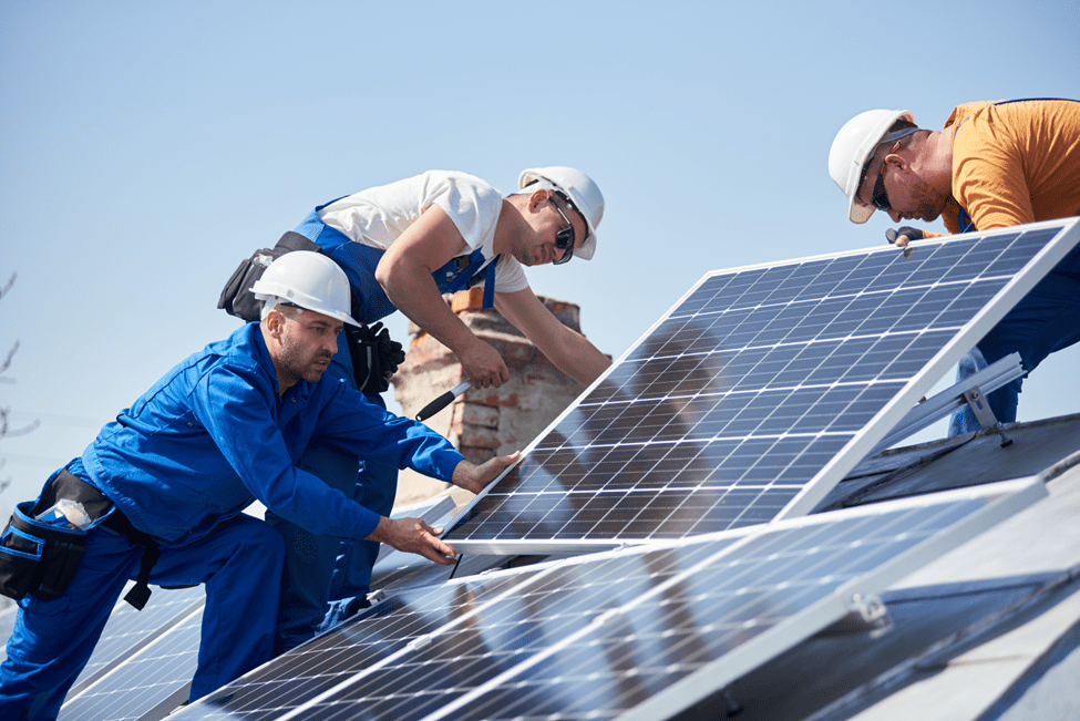 Three workers in safety helmets and uniforms installing solar panels on a sloped rooftop under a clear blue sky.