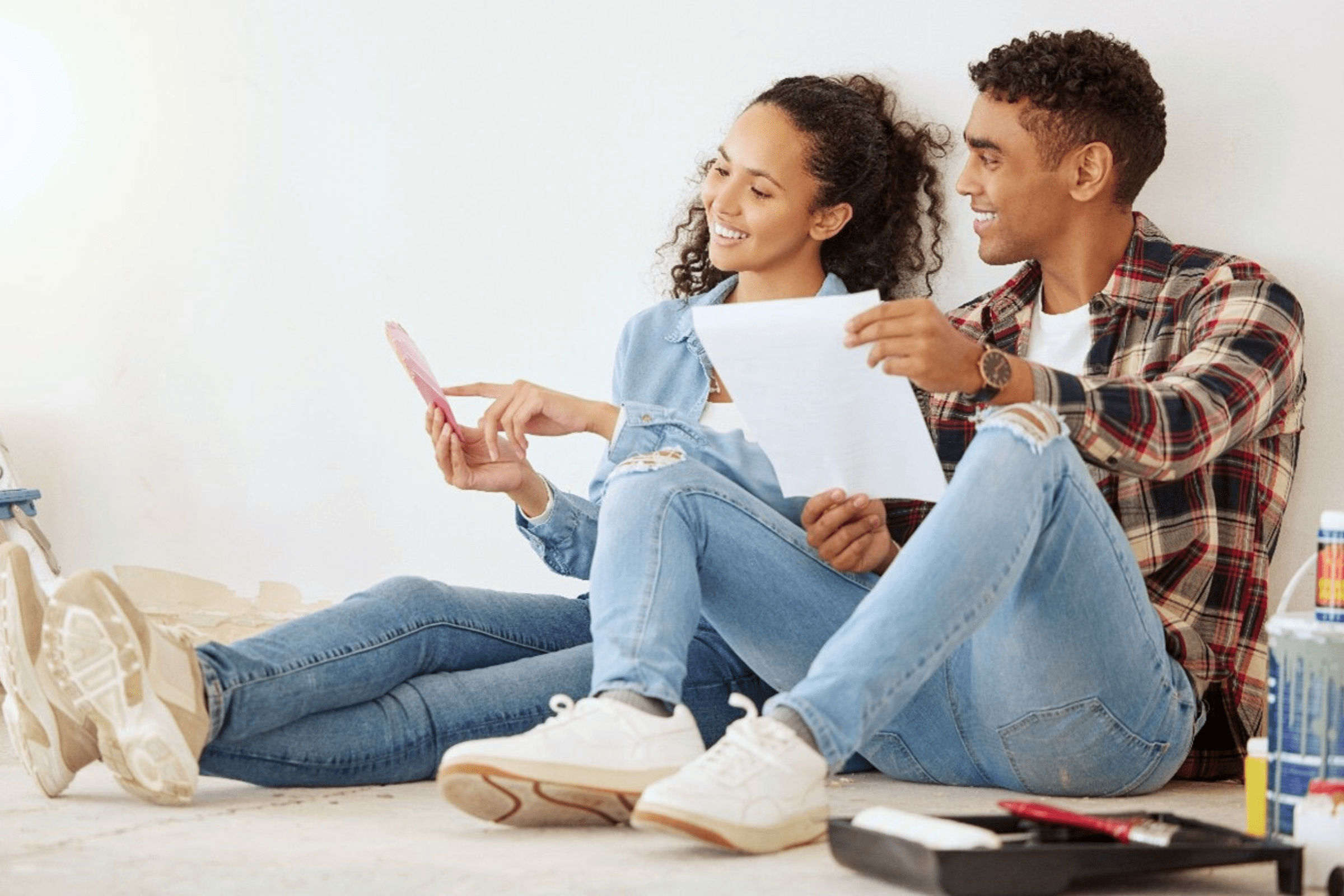 A smiling couple sits on the floor surrounded by paint supplies, discussing renovation plans with a calculator and papers in hand.