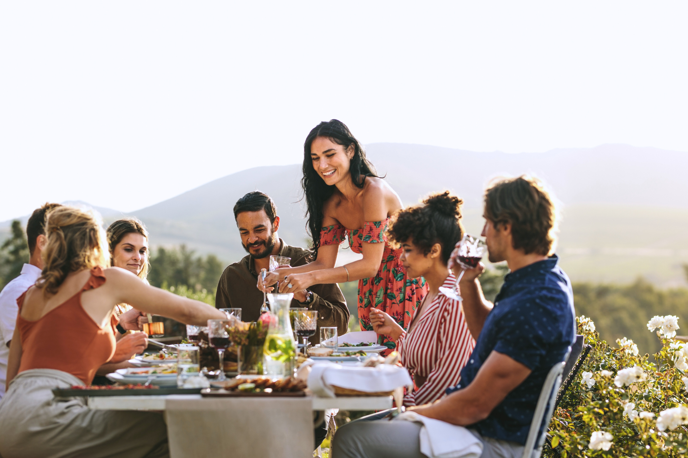 A group of friends having an outdoor dinner party with mountains and flowers in background.