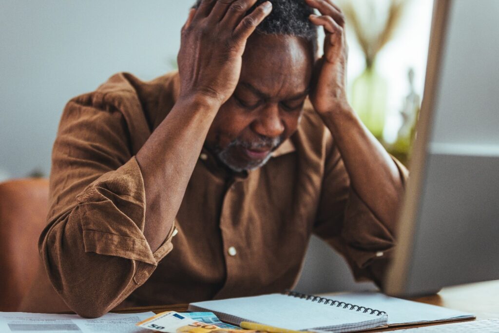 An older man sits at a desk holding his head in frustration while looking at bills, a notebook, and documents.