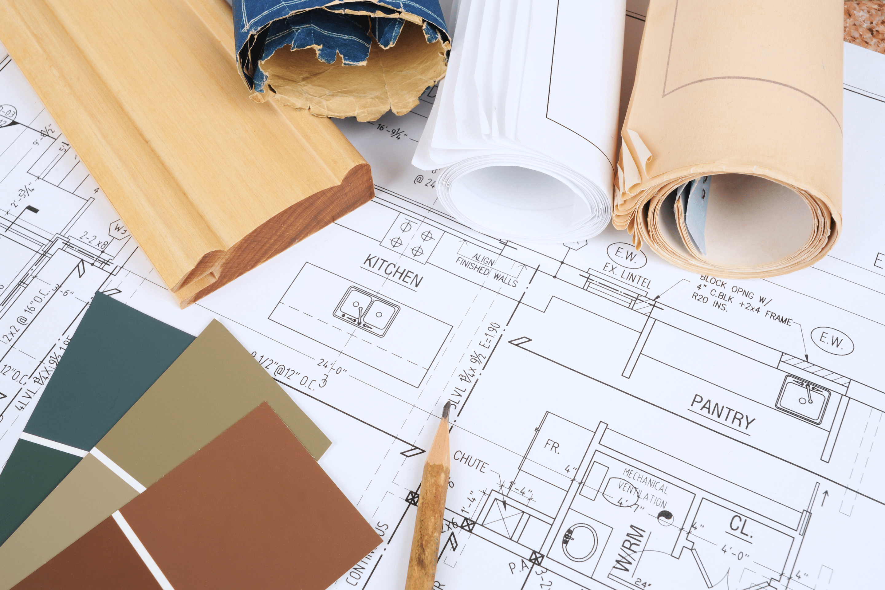 A close-up of home renovation plans showing kitchen and pantry blueprints, surrounded by design materials including paint color swatches, wood trim, rolled architectural drawings, and a pencil.