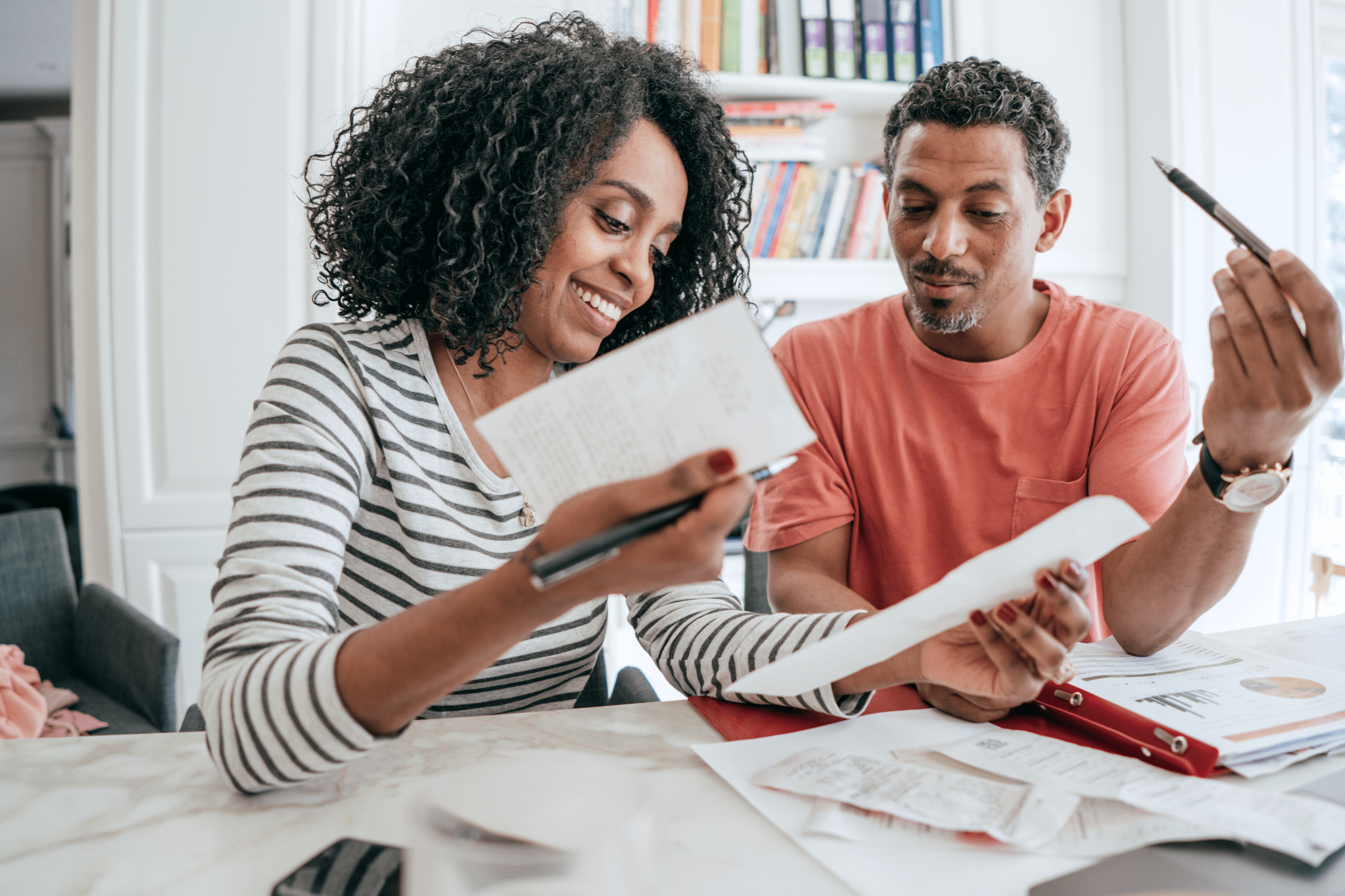 A smiling couple sits at a table reviewing paperwork and receipts. The woman, holding a pen and notebook, looks happy while discussing the documents. The man, also holding a pen, looks engaged in the conversation. Papers and a binder are spread out on the table in front of them.