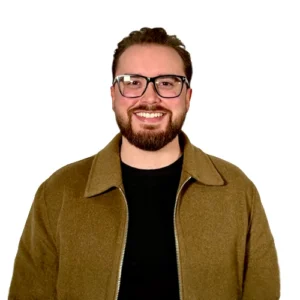 Kyle Smith - WBG - 500x500 Kyle Smith professional headshot wearing glasses, a tan jacket, and a black shirt against a light gray background.
