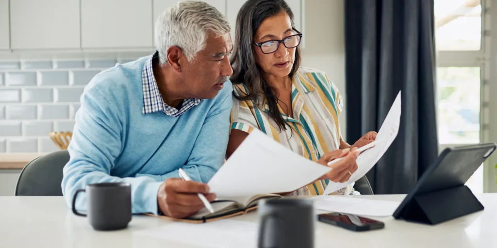 7b20b959-7eff-3632-0696-2f8645fb0653 Two adults reviewing financial documents at a kitchen table with a tablet, notebook, and household budget paperwork.